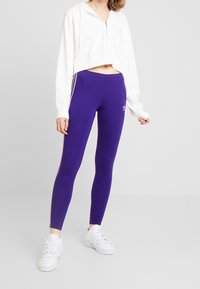 Lila leggings med vita sidosträck, tillsammans med en kortärmad vit hoodie. Leggingsen har en Adidas-logotyp på höften.