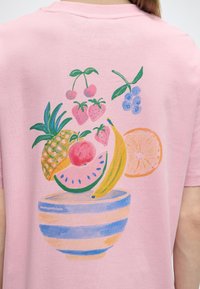 Πίσω μέρος ροζ μπλούζας με πολύχρωμη εικονογράφηση φρούτων: κεράσια, φράουλες, βατόμουρα, ανανάς, ροδάκινο, καρπούζι, μπανάνα, φέτα πορτοκαλιού, σε ριγέ μπολ.