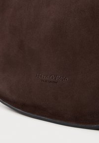 Brązowa torebka z zamszu o miękkiej fakturze, z wytłoczonym logo "Marc O'Polo". Okrągły kształt z czarnym wykończeniem na krawędzi.