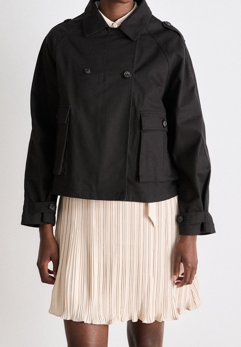 Personne portant une veste noire à double boutonnage avec des poches à rabat boutonnées et une jupe plissée beige sur un fond uni.