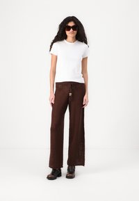 Camiseta blanca de manga corta combinada con pantalones anchos de punto marrones que cuentan con una cinturilla con cordón y acentos de cuentas de madera. Complementado con gafas de sol negras.