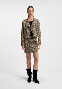 Kvinne som står iført en jakke og skjørt med leopardmønster, med en svart topp og svarte midtlegghøyde hæler mot en hvit bakgrunn.