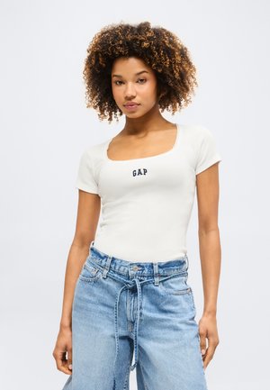 Νεαρή γυναίκα με σγουρά μαλλιά που φοράει λευκό t-shirt GAP και φαρδιά μπλε τζιν με δεμένη ζώνη, στέκεται μπροστά σε ένα μονόχρωμο φόντο.