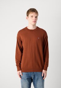 Maglione a maglia marrone con scollatura a girocollo e maniche lunghe, caratterizzato da un logo sottile sul petto. Abbinato a jeans blu sbiaditi.