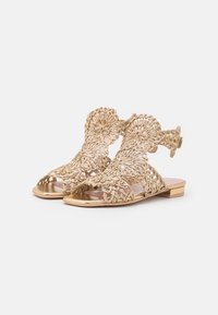 Gul sandal med intrikata blommönster, platt sula, öppen tå och ankelrem. Slät metallisk finish med detaljerad texturerad design.