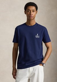 Jonge man die een donkerblauw Polo Ralph Lauren T-shirt en witte broek draagt, staand met één hand in zijn zak tegen een effen achtergrond.