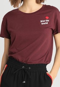 Camiseta de algodón color marrón con cuello redondo y mangas cortas, que presenta el texto blanco "Kiss the world" y un gráfico de labios rojos en el lado izquierdo del pecho.