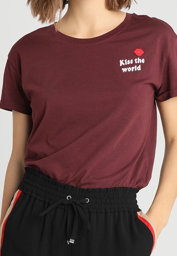 Camiseta de algodón color marrón con cuello redondo y mangas cortas, que presenta el texto blanco "Kiss the world" y un gráfico de labios rojos en el lado izquierdo del pecho.
