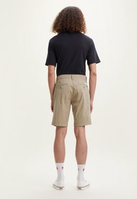 Pantaloni beige con tasche posteriori e una maglietta nera, indossati con calze bianche e sneakers bianche, dal texture liscia e design casual.