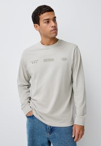 Lysegrå langærmet skjorte med rund halsudskæring, prydet med trykt tekst og ikoner i mørkere nuancer. Kombineret med blå denimjeans.