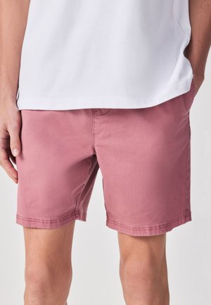 Man die roze korte broek en een wit shirt draagt, staat met één hand in zijn zak tegen een effen achtergrond.