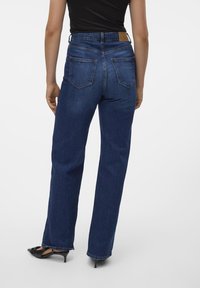 Modri usnjeni jeans visoke pasu in ravnih nogavic, z zadnjimi žepi in rjavim oznako na pasu, v kombinaciji s črnimi čevlji s koničastim nosom.