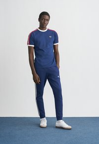 Temno modra oprijeta majica s belim ovratnikom in rdečimi črtami, združena s temnimi trenirkami z belimi bočnimi črtami in logotipom Adidas na prsih.