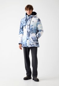 Doudoune à imprimé montagne bleu et blanc, avec capuche, comprenant une fermeture éclair à l'avant et des boutons, des poches latérales et des poignets élastiques.