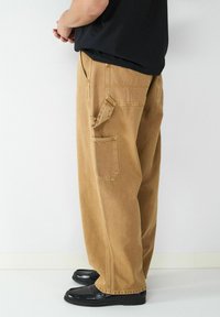 Locker, braune Cordhosen mit mehreren Taschen, gestickten Akzenten und strukturierter Oberfläche, kombiniert mit schwarzen Business-Schuhen.