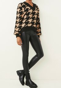 Beżowy i czarny sweter dzianinowy w wzór houndstooth z dekoltem w kształcie litery V, zestawiony z błyszczącymi czarnymi legginsami i masywnymi czarnymi botkami za kostkę.