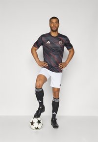 Maillot de football à manches courtes noir et rouge à motifs, avec un logo circulaire, associé à un short blanc et des chaussettes noires, debout sur un ballon de soccer.