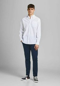 Camicia bianca con bottone e tasca sul petto, abbinata a pantaloni blu navy e scarpe da ginnastica nere con accenti bianchi e rossi. Tessuto dalla consistenza liscia.