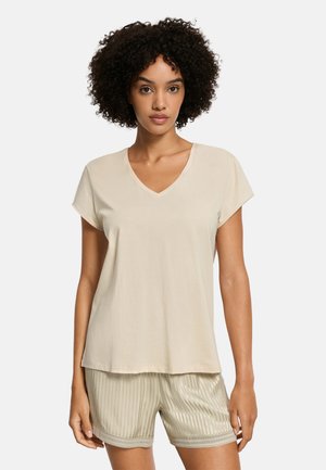 Kobieta o kręconych włosach, ubrana w beżowy top z krótkim rękawem i dekoltem w serek oraz jasne, paskowane szorty na jednolitym tle.