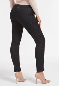 Black patterned pants with a sleek fit, featuring a subtle dot texture, zippered back pocket, and paired with nude high-heeled shoes.