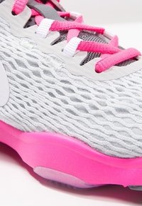 Harmaa urheilukenkä, jossa on teksturoitu verkkopinta, vaaleanpunaisia yksityiskohtia ja paksu, kirkas vaaleanpunainen pohja. Varustettu litteillä nauhoilla ja kaarevalla muotoilulla.