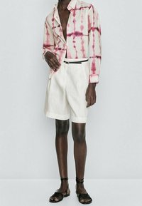 Korte mouwen, tie-dye shirt met roze patronen op een lichte stof, gecombineerd met witte linnen shorts en zwarte sandalen met enkelbanden.