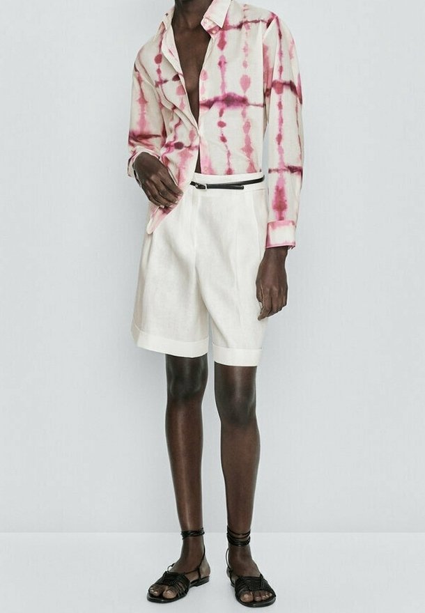 Korte mouwen, tie-dye shirt met roze patronen op een lichte stof, gecombineerd met witte linnen shorts en zwarte sandalen met enkelbanden.