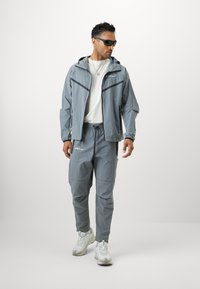 Αθλητική φόρμα σε ανοιχτό γκρι χρώμα, κατασκευασμένη από ελαφρύ υλικό, που περιλαμβάνει ζακέτα με φερμουάρ, παντελόνι με ελαστική μέση και αθλητικά λευκά παπούτσια.