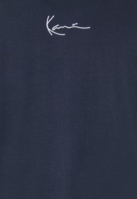 Camiseta azul marino de algodón suave con el texto bordado en blanco "Kanye" que tiene una ligera curvatura. Diseño simple de cuello redondo y textura suave.