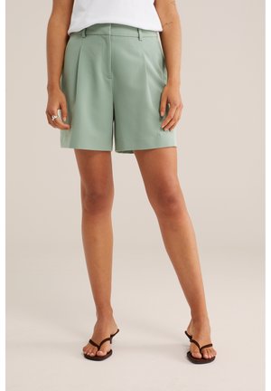 Femme portant un short vert clair sur mesure, un haut blanc et des sandales à talons noires, debout devant un fond uni.