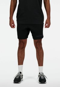 Shorts New Balance noirs avec une texture lisse, arborant un logo sur la jambe gauche. Associés à des baskets noires et des chaussettes blanches.