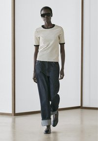 Frau geht drinnen, trägt Sonnenbrille, kurzärmeliges weißes Shirt mit schwarzen Streifen, hochgekrempelte dunkle Jeans und schwarze Schuhe.