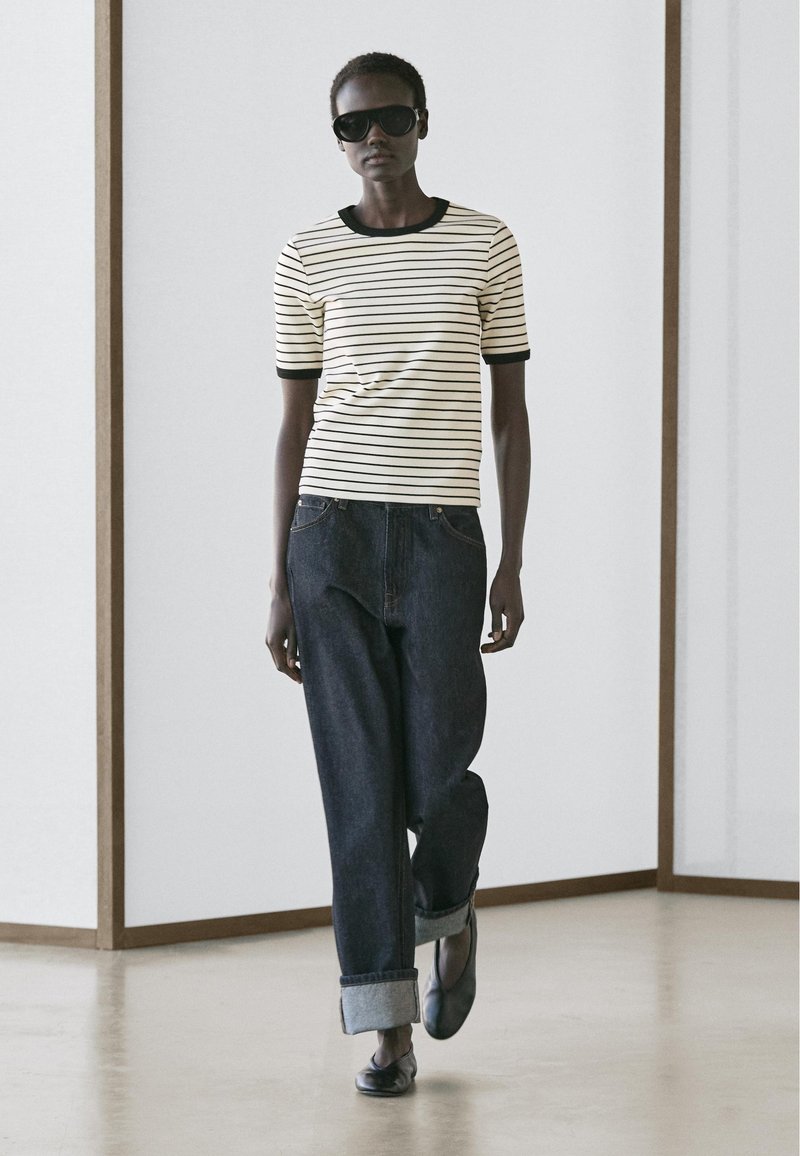 Frau geht drinnen, trägt Sonnenbrille, kurzärmeliges weißes Shirt mit schwarzen Streifen, hochgekrempelte dunkle Jeans und schwarze Schuhe.