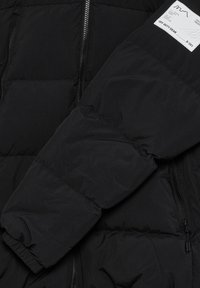 Zwarte pufferjack met een gewatteerd ontwerp, ritssluiting aan de voorkant en elastische manchetten. Bevat een label op de bovenarm met productdetails.