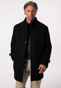 Abrigo de lana negro con un cuello alto, cierre frontal de botones y bolsillos laterales, combinado con una camisa oscura de botones y pantalones claros.