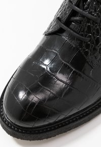 Scarpa in pelle nera con un motivo in pelle di coccodrillo lucido e texturizzato. Presenta una punta arrotondata e lacci neri, su una suola testurizzata in contrasto.