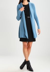 Světle modrý dlouhý kardigan s žebrovanou texturou, s otevřeným předním designem, nošený přes černé šaty, spárovaný s černými kotníkovými botami.
