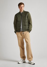 Veste bomber verte olive avec un devant matelassé, pull gris en dessous, pantalon beige et baskets grises avec des accents noirs.