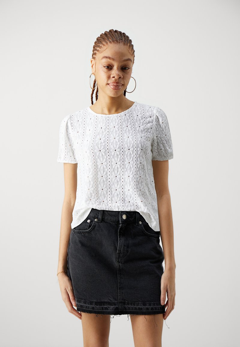 Weißes Kurzarm-Oberteil mit Lochmuster und floralem Design, kombiniert mit einem schwarzen Jeans-Minirock mit ausgefranstem Saum und Fronttaschen.