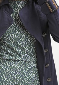 Laivan sininen trench coat, jossa on suuret napit, vyötäröllä vyö ja leveä kaulus, on kerroksittain vihreän kuviollisen mekon päällä, jossa on pieniä valkoisia ja sinisiä pisteitä.