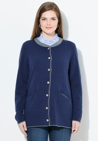 Vrouw in een marineblauwe ge-button cardigan met grijze afwerking, een blauw geblokte blouse met een gerimpelde kraag en spijkerbroek, staande tegen een witte achtergrond.