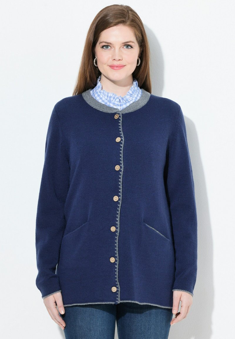 Vrouw in een marineblauwe ge-button cardigan met grijze afwerking, een blauw geblokte blouse met een gerimpelde kraag en spijkerbroek, staande tegen een witte achtergrond.