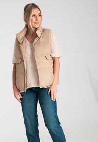 Beżowa kamizelka puchowa z wysokim kołnierzem i zapięciem na napy z przodu, noszona na jasnym swetrze dzianinowym i ciemnoniebieskich dżinsach, z bocznymi kieszeniami.