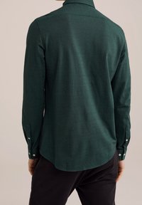 Mörkgrön långärmad skjorta med en slät textur, rundad kant och knappade manschetter. Har en klassisk krage och en figurnära design.