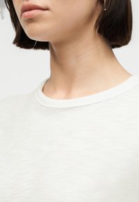 Femme aux cheveux courts et foncés portant un t-shirt blanc à col ras du cou, visible du menton à la poitrine, sur un fond clair.