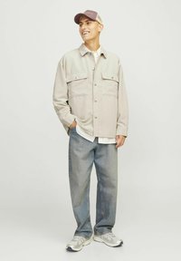 Light beige corduroy shirt jacket with two front pockets, paired with a white shirt and loose blue jeans. Gray sneakers and a light brown cap.