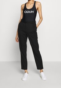 Bañador negro de una pieza con el logo blanco "HUGO", combinado con pantalones negros ajustados y zapatillas blancas, mostrando un conjunto elegante y moderno.