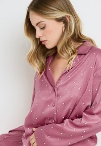 Haut de pyjama en satin rose avec un col en V, orné de petits motifs de cœurs blancs et d'une fermeture à boutons, à manches longues avec une coupe décontractée.