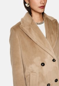 Beige dubbelzijdig jasje met een zachte, pluizige textuur, oversized revers, zwarte knopen en zijzakken. Geaccentueerd met een statement ketting.