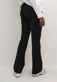 Pantalons noirs évasés en tissu extensible, avec une texture lisse et une taille haute, associés à des baskets blanches avec un design subtil.